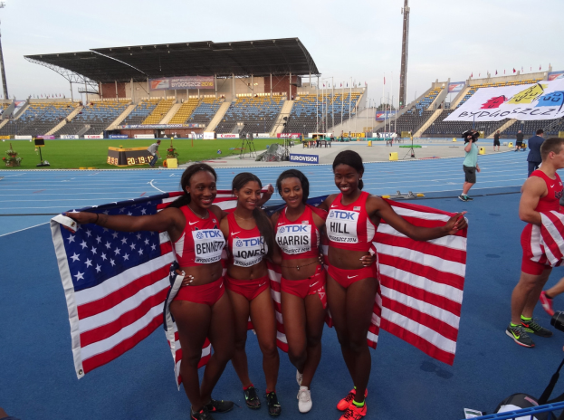 Vier Frauen in Sportkleidung stehen zusammen auf einer Laufbahn, jede hält eine amerikanische Flagge und strahlt, mit einem Stadion und Zuschauern im Hintergrund.
