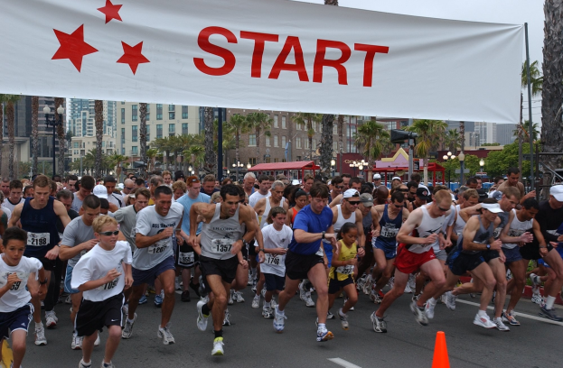 Eine Gruppe von Läufern bei einem Marathon mit einem Verkehrskegel im Vordergrund und einer Fahne im Hintergrund, vor einer Kulisse aus Bäumen, Laternenmasten, Gebäuden und einem klaren blauen Himmel.