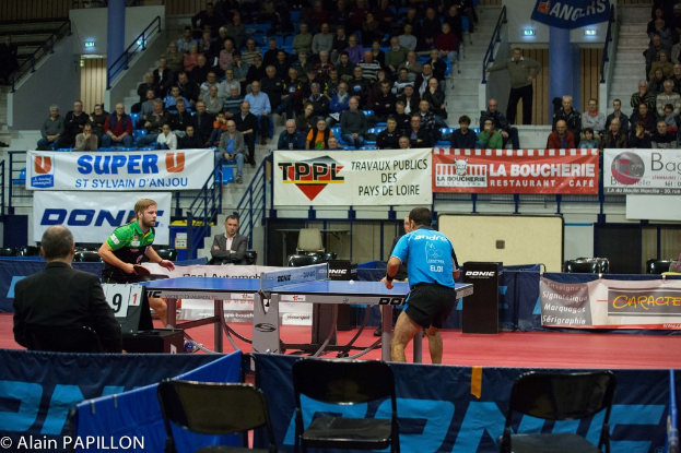 Zwei Männer spielen Tischtennis drinnen, mit mehreren Zuschauern, die stehen und sitzen, einigen leeren Stühlen und Werbetafeln, mit Text in der unteren linken Ecke.