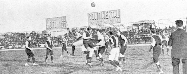 Schwarzes und weißes Foto von Männern, die Fußball spielen, mit einem Ball in der Luft, Zuschauern im Hintergrund und Infotafeln.