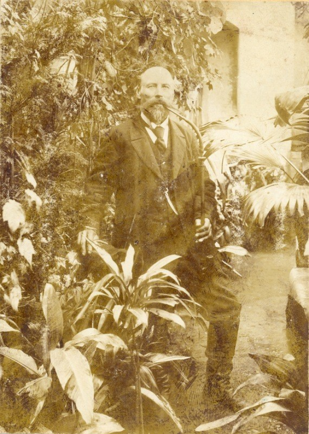 Altes Foto eines Mannes in Anzug und Krawatte, der in einem Garten mit Pflanzen und Bäumen steht, mit einer Wand im Hintergrund.