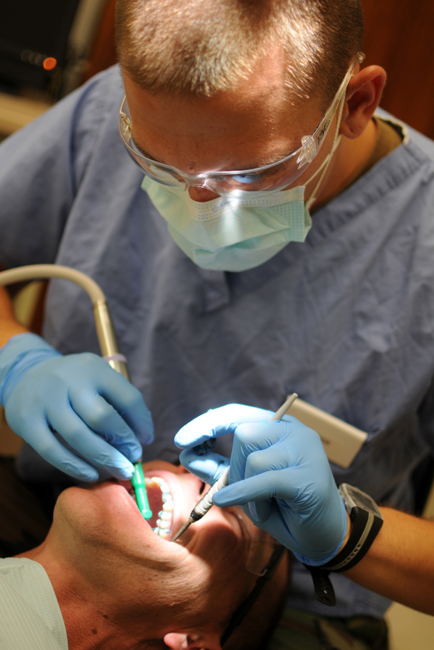 Ein Zahnarzt in Brille, Mundschutz und Handschuhen führt eine zahnärztliche Behandlung an einem Patienten durch, der in einem Stuhl liegt, mit einem unscharfen Hintergrund.