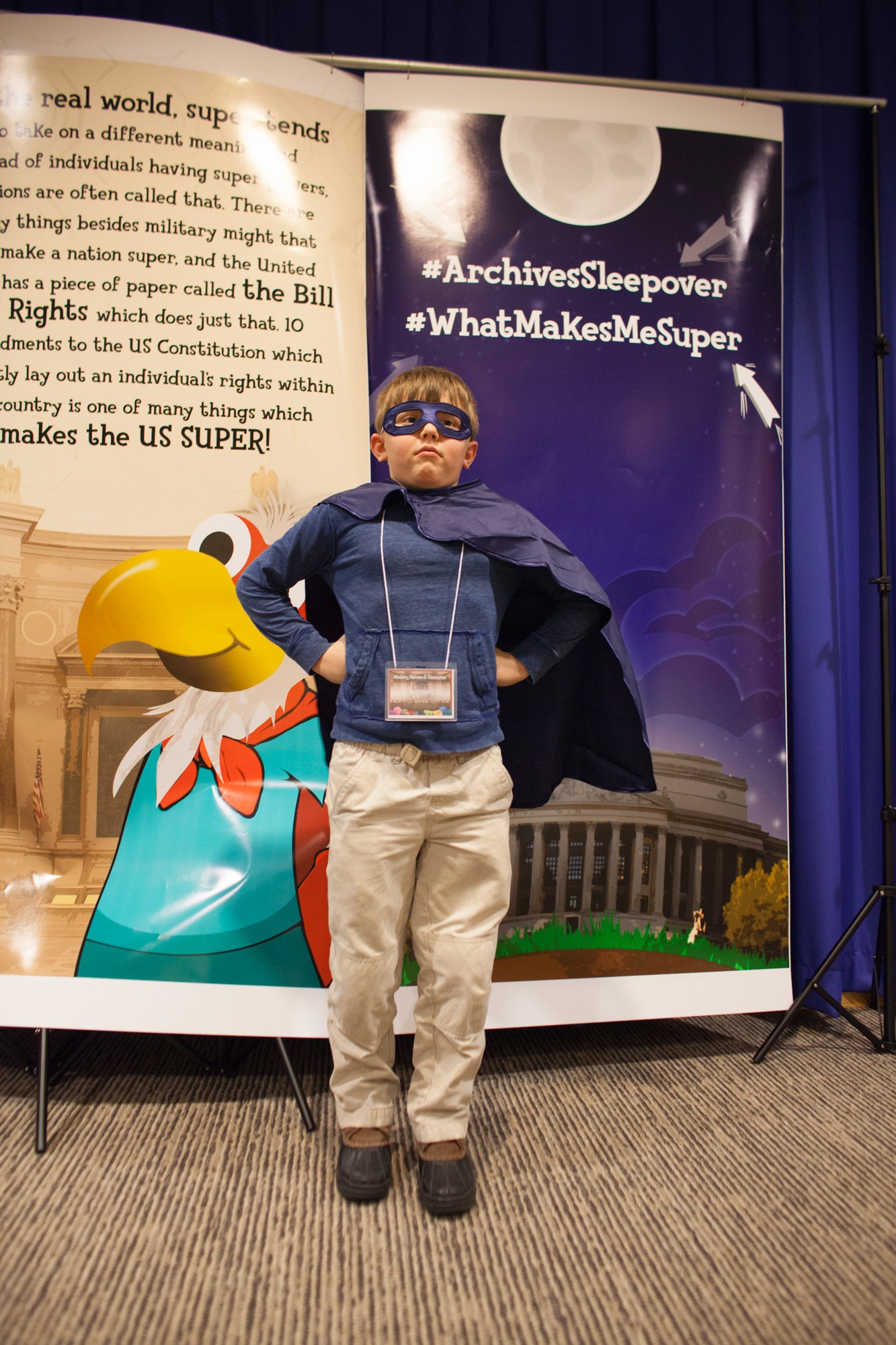 Ein junger Junge in einem Superheldenkostüm steht vor einer Fahne mit Text und einem Vogelbild, mit einem Vorhang im Hintergrund und Schuhen.