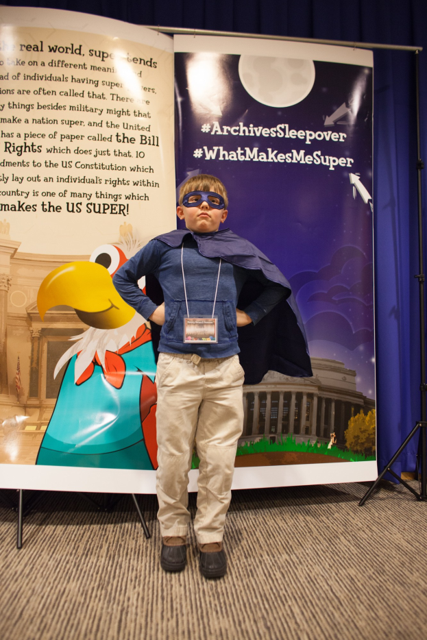 Ein junger Junge in einem Superheldenkostüm steht vor einer Fahne mit Text und einem Vogelbild, mit einem Vorhang im Hintergrund und Schuhen.