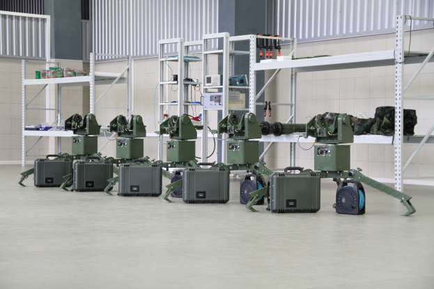 Eine Gruppe von Militärrobotern auf einem Boden mit Koffern und Gegenständen auf Gestellen, vor einer Wand mit Geländer.