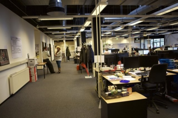 Großer offener Büroraum mit Schreibtischen, Stühlen, Computern, Monitoren, Tastaturen, Papieren, Büchern, Kartons, hängenden Kleidungsstücken, wandmontierten Tafeln mit Text und Bildern, Deckenleuchten und Glasfenstern im Hintergrund.