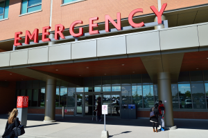 Eine Gruppe von Menschen vor einem Glaswand-Notfallgebäude mit Säulen, einem Namensschild, Schildern auf Ständern und einem Mülleimer.