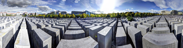 Luftaufnahme des Holocaust-Mahnmals für die ermordeten Juden Europas in Berlin, Deutschland, mit einem Gitternetz aus Betonsteinen, umgeben von Bäumen und städtischen Gebäuden, mit Fahrzeugen auf einer nahen Straße und einem bewölkten Himmel.