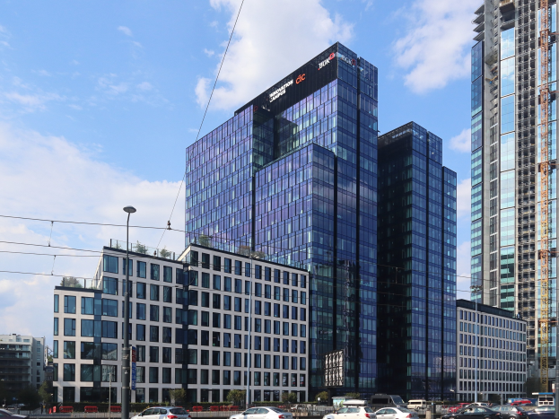 Großes modernes Bürogebäude mit zahlreichen Fenstern, identifiziert als Deutsche Bank Hauptsitz in Frankfurt, Deutschland, umgeben von städtischer Infrastruktur einschließlich Straßenlaternen, Fahrzeugen, Bäumen und einem bewölkten Himmel.