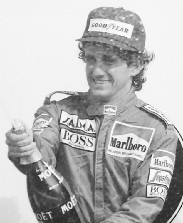 Ayrton Senna, ein Formel-1-Fahrer, steht lachend in einem Rennanzug und einer Mütze, mit einer Flasche Champagner in der Hand.