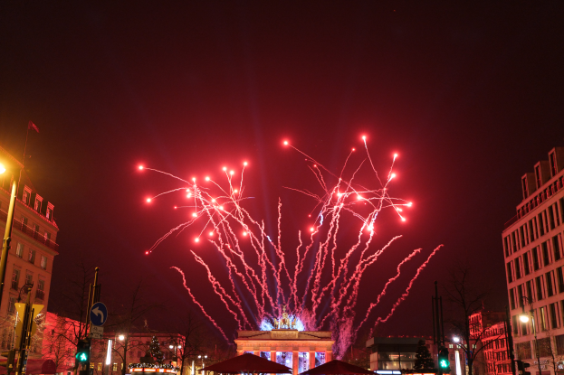 Eine Straßenansicht in Berlin an Silvester, voller Gebäude, Bäume, Laternenpfähle, Verkehrszeichen, Zelte und Menschen, mit einem Feuerwerk erleuchteten Himmel im Hintergrund.