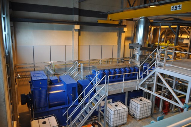 Ein Fabrikinnenraum mit Trägern, einer Treppe mit Geländer, Containern, Deckenleuchten, Rohren und Wänden.