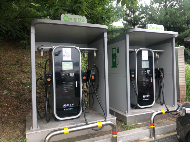 Zwei Elektroauto-Ladestationen nebeneinander, umgeben von Bäumen unter einem klaren blauen Himmel, mit einem geparkten Auto auf der rechten Seite und anderen Objekten auf der linken Seite in Seoul, Südkorea.