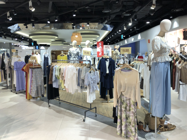 Ein hell erleuchteter Bekleidungsladen mit Ständern voller Kleider, Schaufensterpuppen in Outfits, Informationsschildern, Deckenleuchten und verstreuten Gegenständen auf dem Boden.