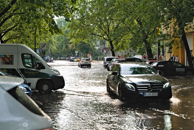 Eine überflutete Straße in Berlin, Deutschland, mit Autos, die durch das Wasser fahren, Menschen, die auf dem Gehweg gehen, Bäume, die die Straße säumen, Gebäude im Hintergrund und Schilder und Pfosten im Wasser, unter einem sichtbaren Himmel.