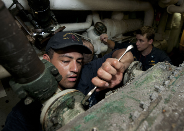 Zwei Männer in einer Fabrik, die an einer Maschine arbeiten, einer hält ein Werkzeug, mit Rohren und Equipment im Hintergrund.