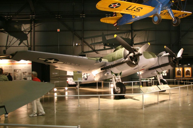 Ein Innenblick in eine Halle mit Flugzeugen, eines hängt von der Decke, Lampen auf dem Boden, Menschen in der Nähe eines Zauns und eine Tafel mit Text an der Wand, beleuchtet von nahen Lampen.