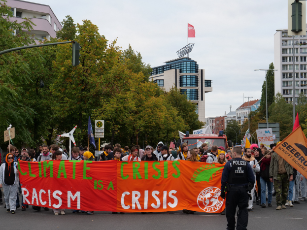 Eine Gruppe von Menschen marschiert auf einer von Bäumen gesäumten Straße, die eine Fahne schwingt, auf der steht "Klima-Krise ist eine Krise", mit Gebäuden und einem klaren blauen Himmel im Hintergrund.