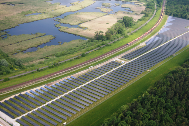 Eine Luftaufnahme eines Solarparks mit Panelen in einem Feld, umgeben von Bäumen, Gras, Pflanzen und Wasser, mit einem Zug auf einem nahen Bahngeleis.