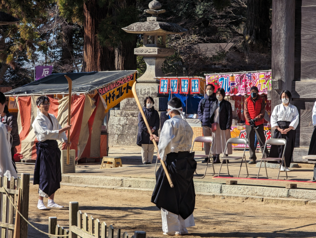 Eine Gruppe von Menschen in traditioneller Kleidung versammelt sich im Freien in Kyoto, einige tragen Masken und halten Holzstöcke, mit Bannern, einem Zelt und einem Holzzaun gegen einen klaren blauen Himmel.