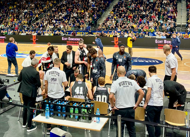 Eine Gruppe von Männern steht auf einem Basketballfeld umgeben von Zuschauern, mit einer Person, die eine Kamera hält, auf der linken Seite und Tafeln mit Text im Hintergrund.