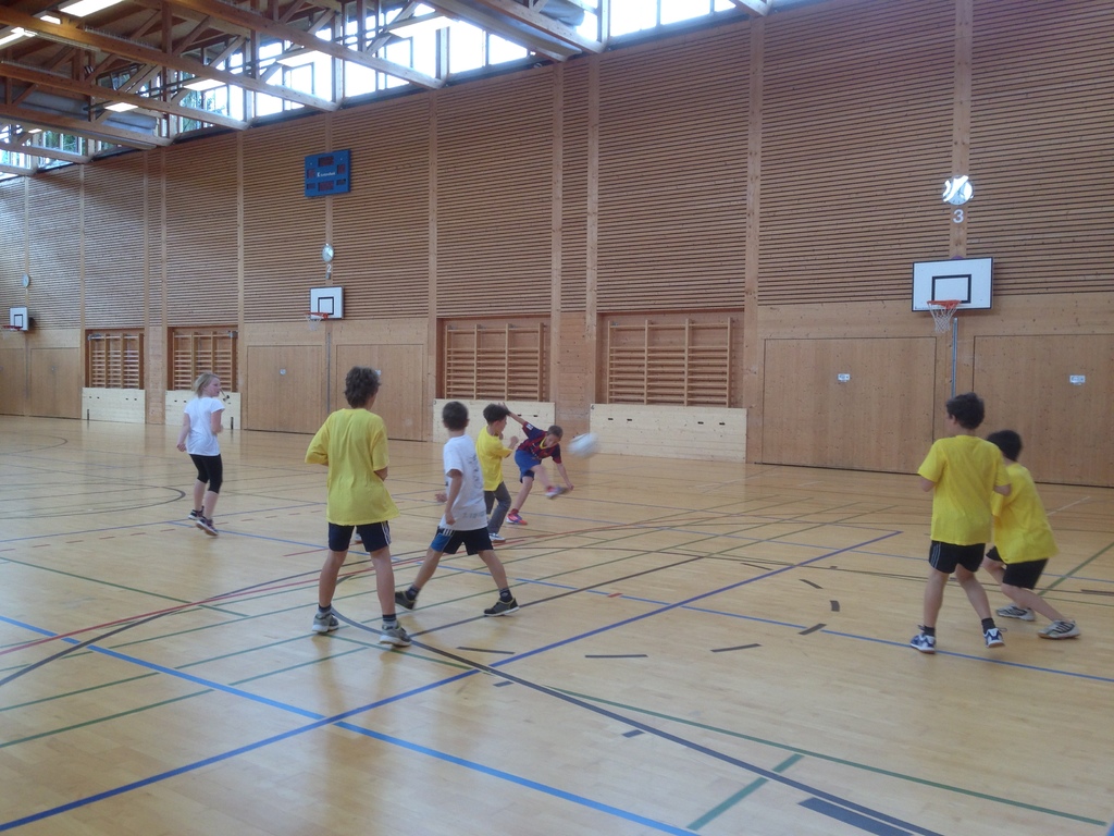 Kinder spielen mit einem Ball vor einer hölzernen Wand mit Basketballkörben, hölzernen Stangen und Fenstern oben.