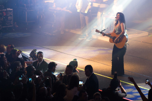 Eine Gruppe von Menschen auf der Bühne, die Musikinstrumente spielen und singen, mit sichtbaren elektrischen Geräten. Im Publikum halten einige Menschen Mobiltelefone und eine Person eine Kamera.