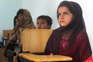Eine Gruppe von Kindern sitzt an Tischen in einem Klassenzimmer in Afghanistan und nimmt an einem Unterricht teil, mit einer Wand im Hintergrund.