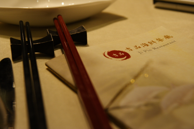 Ein Tisch mit einer Speisekarte, die "Ji Pin Restaurant" auf Mandarin und Englisch lautet, sowie zwei Paar Stäbchen (eines rot, eines schwarz) und eine weiße Schüssel vor den Stäbchen.