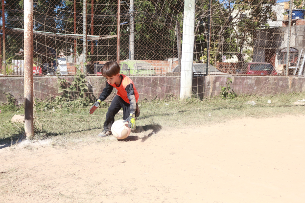 Ein junger Junge spielt Fußball auf einem Schotterfeld mit Gras, Pflanzen, Pfählen, einem Zaun, einer Wand, Bäumen, Fahrzeugen, Gebäuden und einem bewölkten Himmel im Hintergrund, trägt Schuhe und ist aktiv im Spiel.
