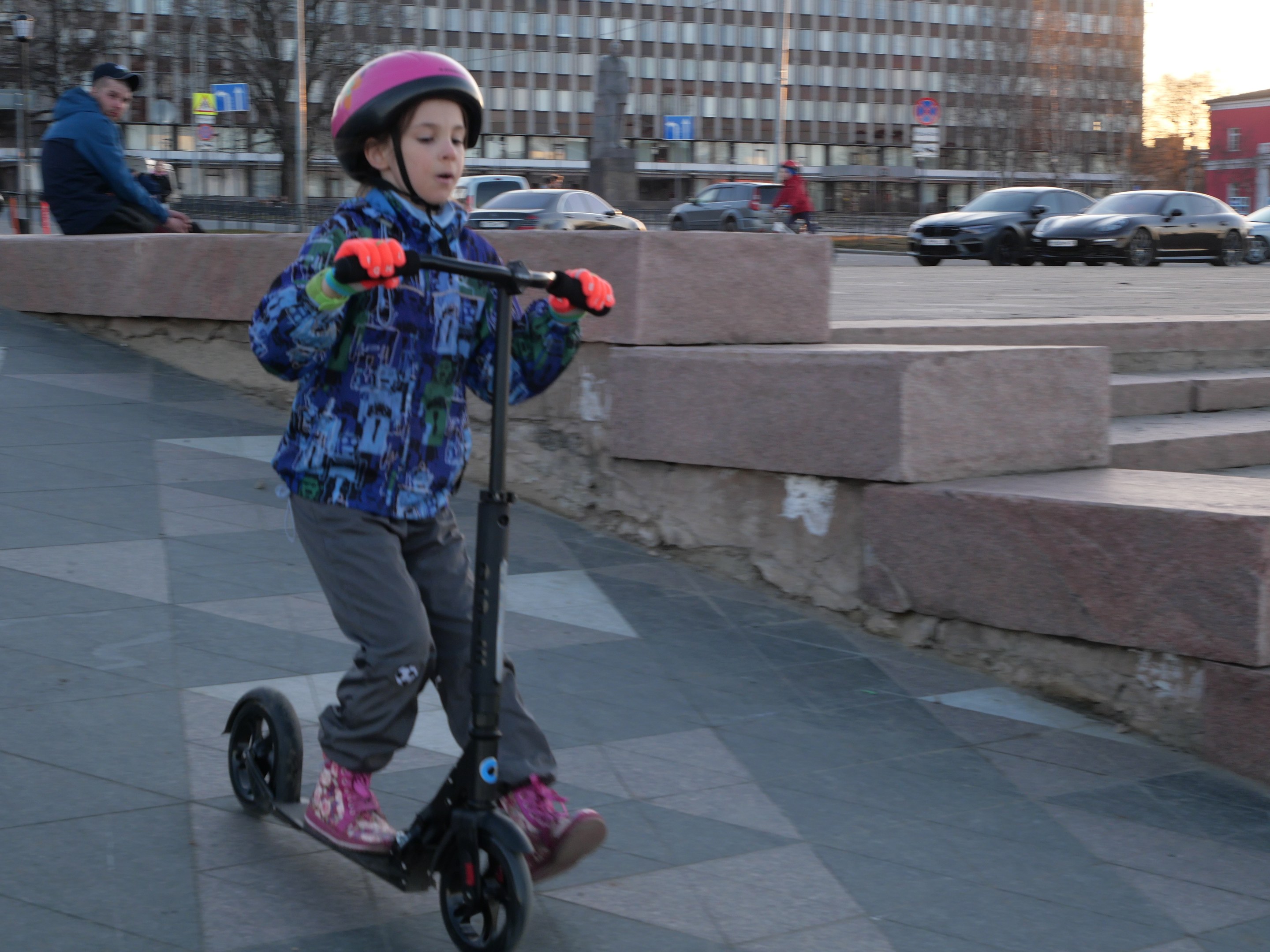 Ein junger Junge in einem Helm und Handschuhen fährt einen Roller eine Treppe hinunter, vorbei an Fahrzeugen, Menschen, Bäumen, Pfosten, Brettern, Gebäuden und einem klaren blauen Himmel im Hintergrund.