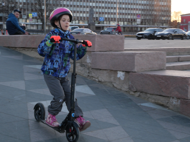 Ein junger Junge in einem Helm und Handschuhen fährt einen Roller eine Treppe hinunter, vorbei an Fahrzeugen, Menschen, Bäumen, Pfosten, Brettern, Gebäuden und einem klaren blauen Himmel im Hintergrund.