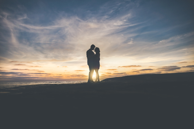 Silhouette eines Paares, das sich auf einem Strand bei Sonnenuntergang umarmt und küsst, mit leuchtendem Orange, Pink und Purpurhimmel, tiefblauem Wasser und der untergehenden Sonne im Hintergrund.