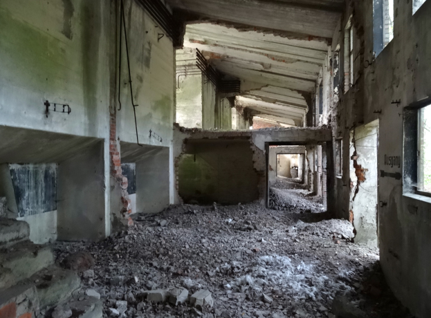 Ein altes, baufälliges Gebäude mit kaputten Fenstern, verstreutem Schutt und verschiedenen Gegenständen wie Rohren und Trümmern auf dem Boden.