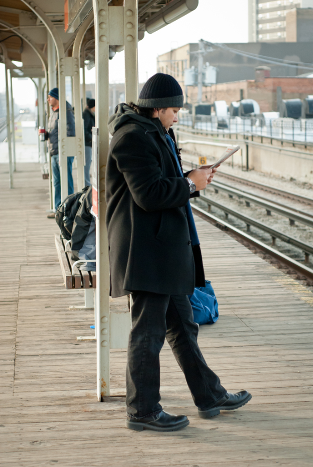 Ein Mann in einer schwarzen Jacke steht an einem Bahnhof mit einem Eisenpfahl und einem Schuppen dahinter und einer Bahnlinie mit braunen Gebäuden auf der rechten Seite.