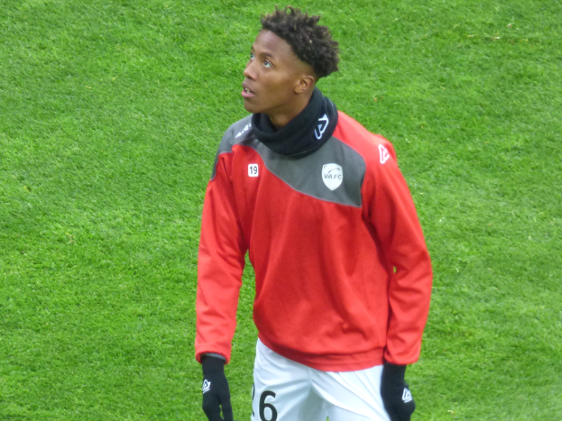 Ein junger Mann in einer roten Jacke und weißen Shorts steht auf einem Fußballfeld und schaut rechts mit entschlossener Miene, trägt Baseballhandschuhe.