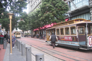 Ein Zug auf Schienen mit Text an der Seite, eine Person in der Nähe, kleine Pfosten mit Seilen auf der linken Seite, Straßenlaternen, Bäume, viele Menschen im Hintergrund, ein Müllcontainer auf der linken Seite und Gebäude mit Leitern auf der rechten Seite.