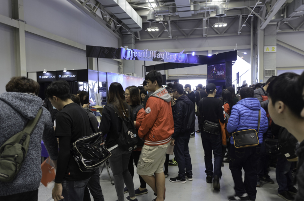 Eine Gruppe von Menschen steht vor einer Menge auf einer Gaming-Messe, wobei einige Besucher Taschen, Texttafeln, Deckenleuchten und Rohre im Hintergrund tragen.