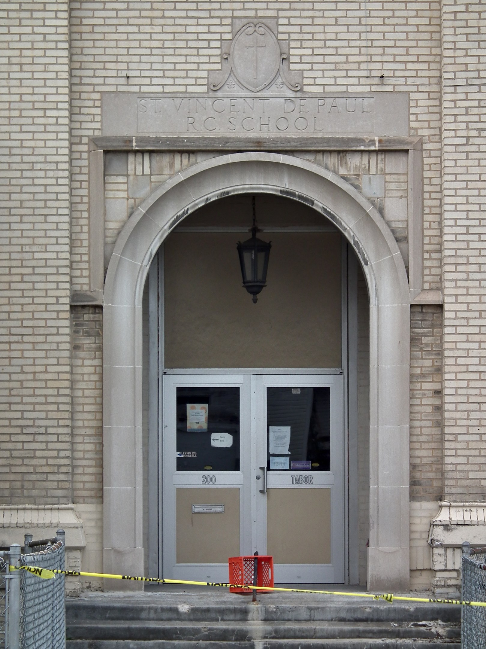 Eingang zu einem Backsteingebäude mit gelbem Absperrband, Treppe mit Maschendrahtzaun, eine Tür mit Papieren, eine hängende Lampe und ein Name an der Wand.