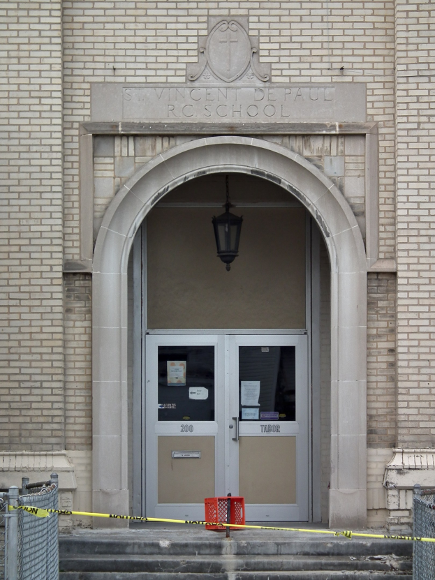 Eingang zu einem Backsteingebäude mit gelbem Absperrband, Treppe mit Maschendrahtzaun, eine Tür mit Papieren, eine hängende Lampe und ein Name an der Wand.