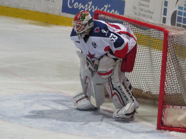 Ein Hockey-Torwart in voller Ausrüstung steht in seinem Netz auf dem Eis, mit einer textbedeckten Wand im Hintergrund.