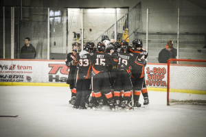 Eine Gruppe von Menschen in Schlittschuhen, Handschuhen und Helmen steht auf einem Eisboden mit Absperrungen und Glastüren dahinter, ein Hockey-Tor mit Netz auf der rechten Seite und ein Hockey-Schläger auf der linken Seite.