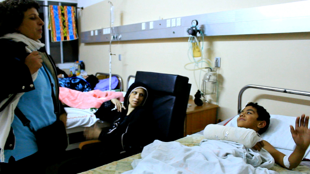 Ein Krankenhauszimmer mit mehreren Betten, auf denen Patienten liegen, und Infusionsflaschen auf Stönden zwischen den Betten, sowie eine Frau mit einer Tasche in der Nühhe eines Patienten.