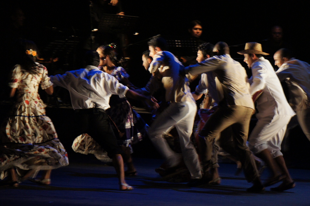 Eine Gruppe von Menschen tanzt im Vordergrund mit einer anderen Gruppe, Stühle und Mikrofone im Hintergrund, alles auf einem Boden.