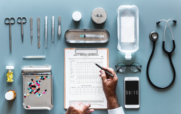 Ein Arzt schreibt auf einem Clipboard mit einem Stethoskop neben verschiedenen medizinischen Hilfsmitteln, darunter ein Mobiltelefon, eine Brille, eine Flasche, eine Schere, eine Spritze, eine Pillenschachtel und andere Gegenstände auf einer blauen Oberfläche.