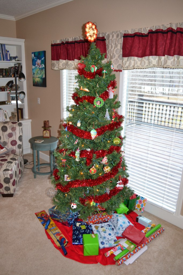 Ein Weihnachtsbaum mit Geschenken davor, eine Couch, ein Tisch, ein Bücherregal, ein Wandbild und eine Stehlampe mit Vorhängen im Hintergrund.