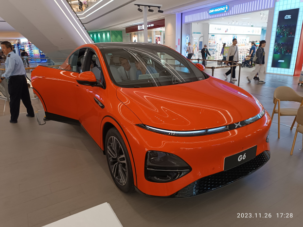 Oranger Elektroauto in einer Ausstellungshalle mit Menschen drumherum, umgeben von Stühlen, einem Wagen, Geschäften, Namensschildern und Lichtern im Hintergrund.