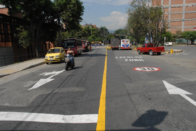 Eine Straßenszene mit Gebäuden, Bäumen, fahrenden Fahrzeugen, Verkehrsschildern, Ampeln, Warnmarkierungen und einem blauen bewölkten Himmel.
