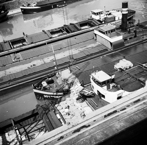 Ein Schwarz-Weiß-Foto eines festgemachten Schiffs in ruhigem Wasser, mit anderen Booten im Hintergrund und einer umgebenden Wand.