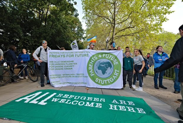 Eine Gruppe von Menschen, die draußen vor einem Banner mit der Aufschrift "Alle Flüchtlinge sind willkommen" und einer Flagge stehen und Fahrräder, ein Zaun, ein Straßenpfahl, ein Schild, Bäume und einen bewölkten Himmel im Hintergrund sehen.